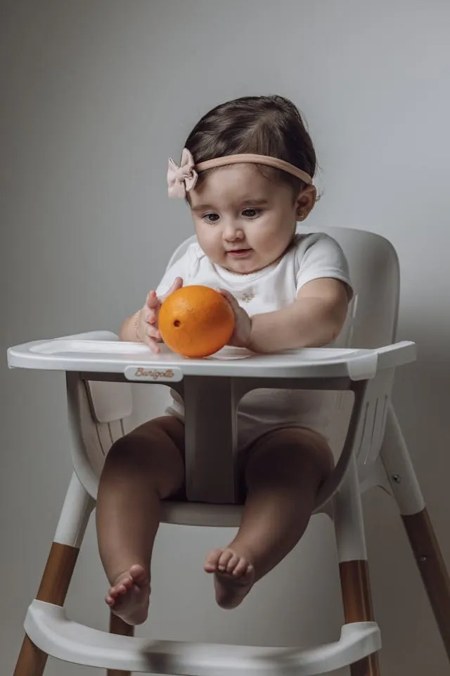 A baby in a high chair holding an orange with both hands
